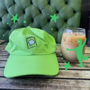 Bright green baseball cap on a worn wooden table. The Crucial Comix logo, a doodle of a zine, is embroidered on the front of the hat.