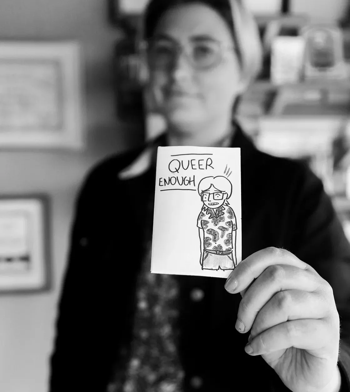 A black and white portrait shows someone holding up a zine called Queer Enough