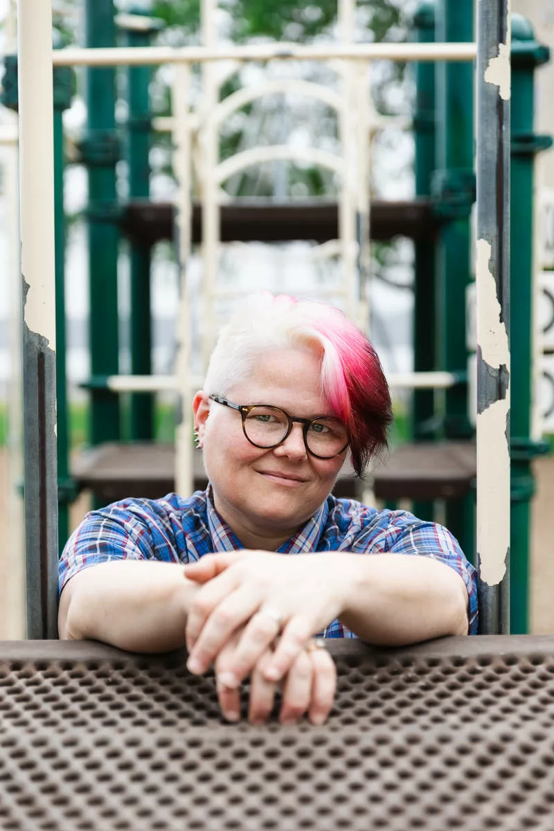 Photo of Sara Ryan, who has light skin and short, white hair with a vibrant shock of red at the front. They wear dark glasses and a blue plaid shirt. Their hands are clasped in front of them as they look toward the camera.